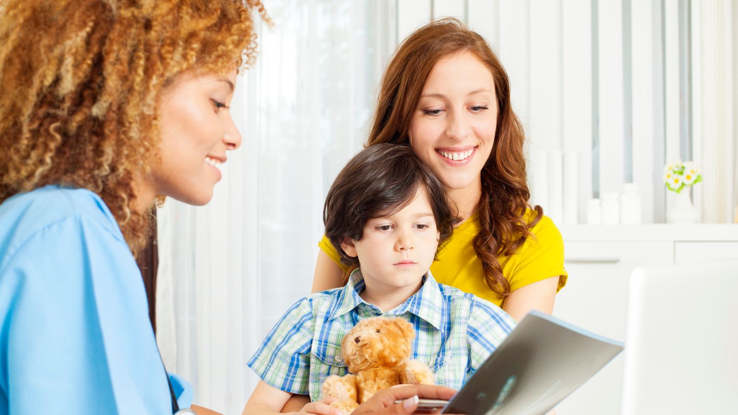 Little boy holding teddy bear, sitting on mothers lap and examining his hand x-ray image together with doctor and his mother.