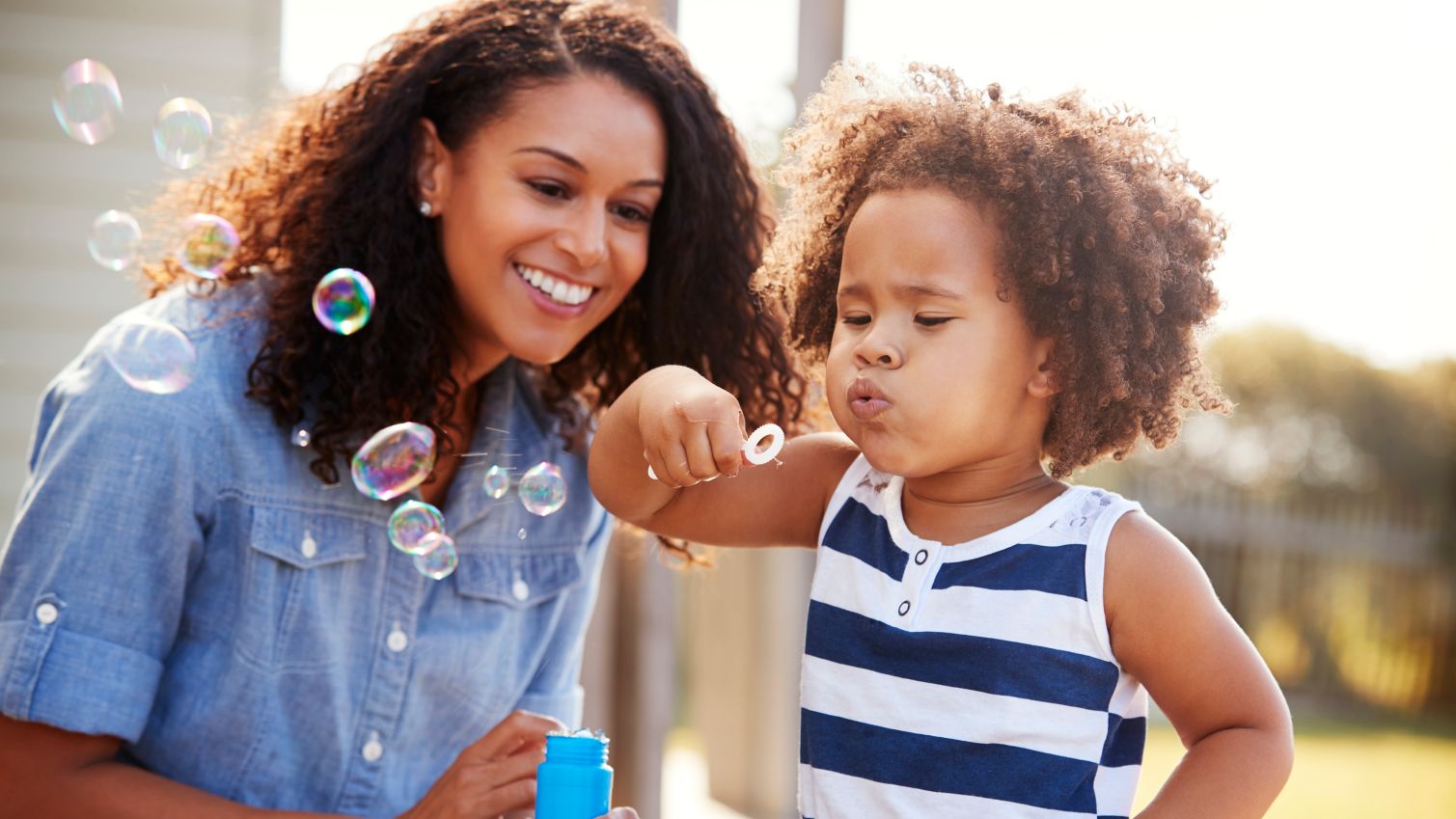 Young child blows bubbles outside while mother looks on