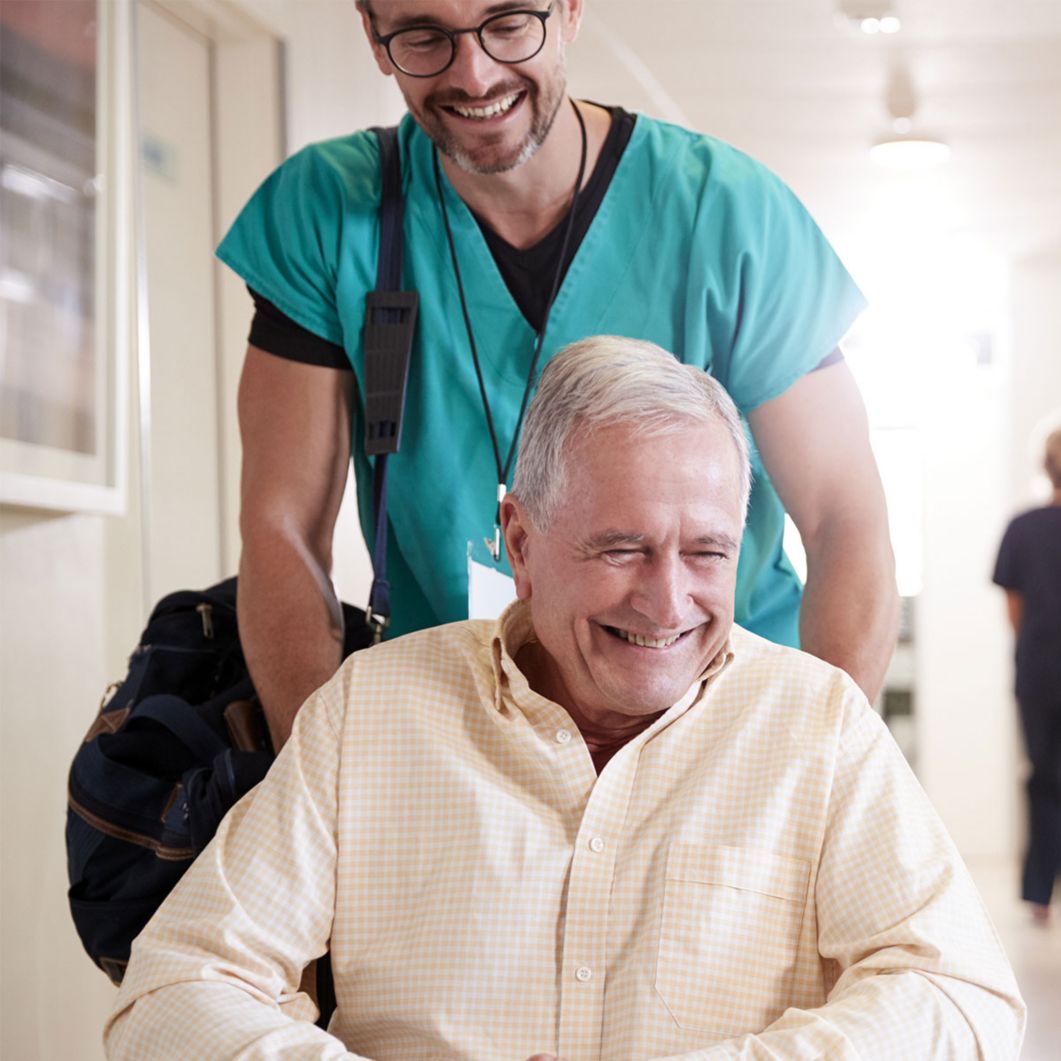 A man in a wheelchair smiles while talking to the caregiver pushing him. 