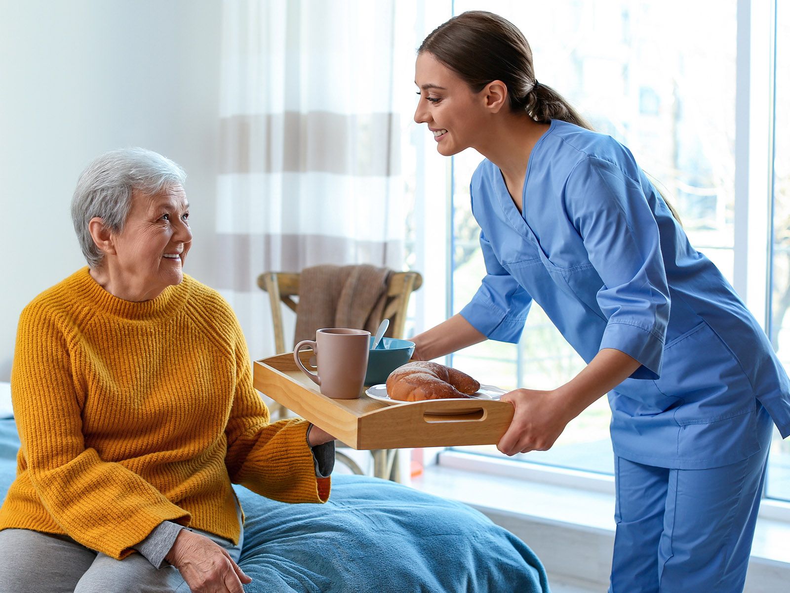 Young female nurse delivers meal to older female patient sitting on bed.