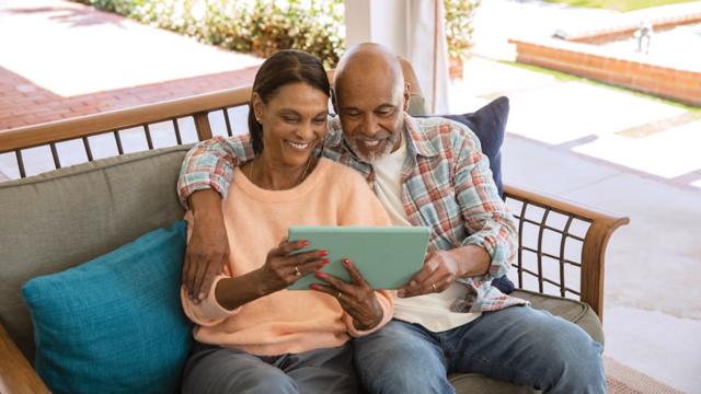 A couple smiles while sitting on their patio with a tablet.