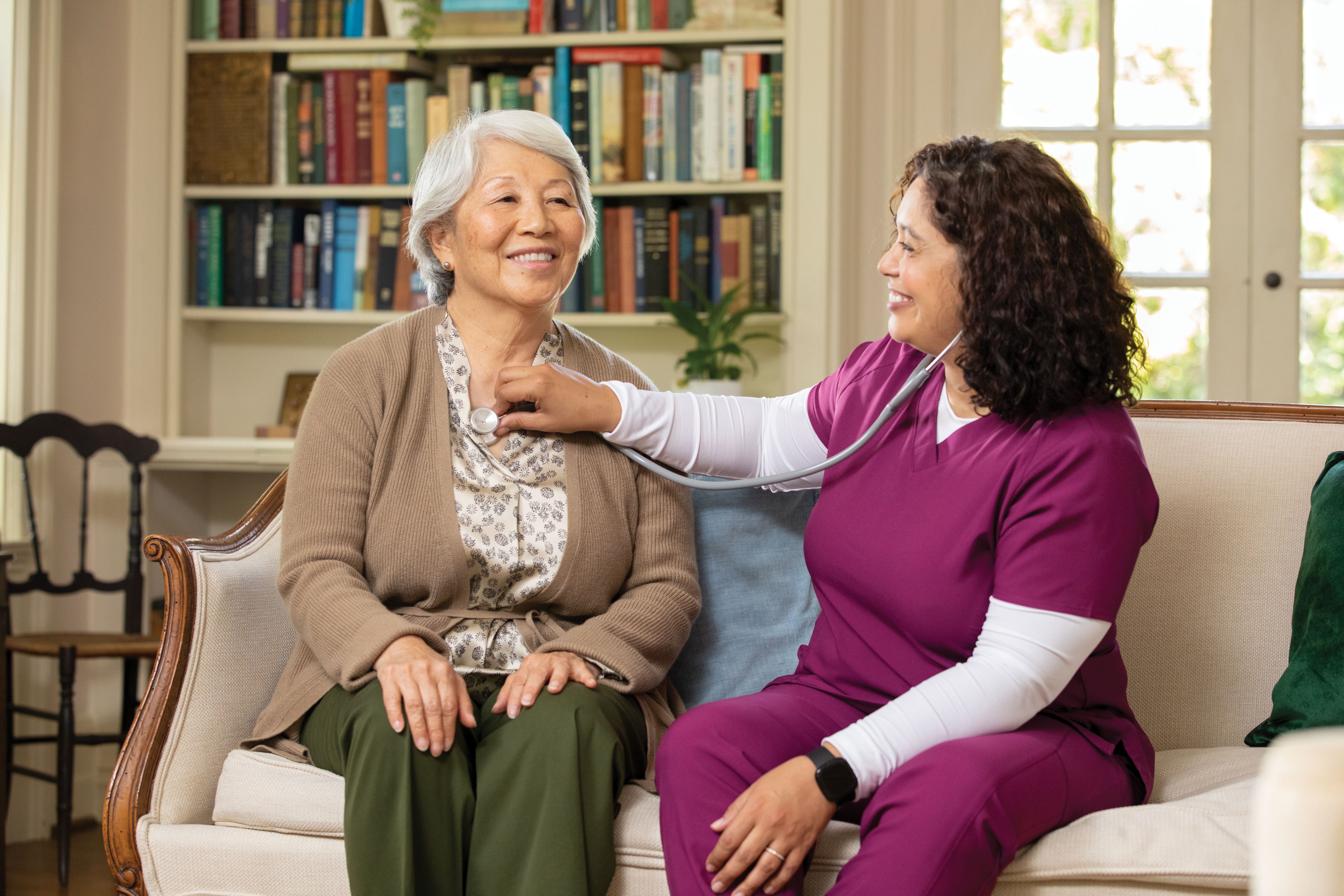 Senior woman sits on couch getting a checkup with caregiver using a stethoscope