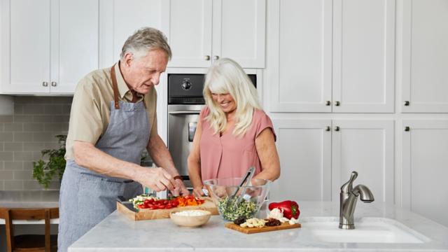 A couple laughs while chopping vegetables in their kitchen.