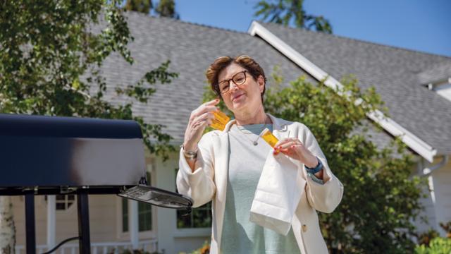 A woman stands at her mailbox and reads the labels on prescription bottles she's pulled from a package.