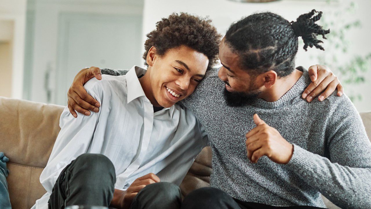 Young man and older man hugging in support while sitting on a couch