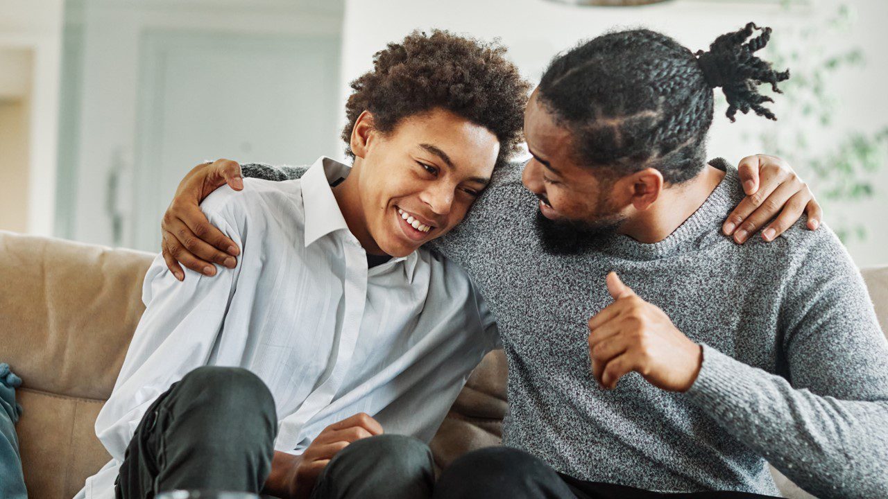 Young man and older man hugging in support while sitting on a couch