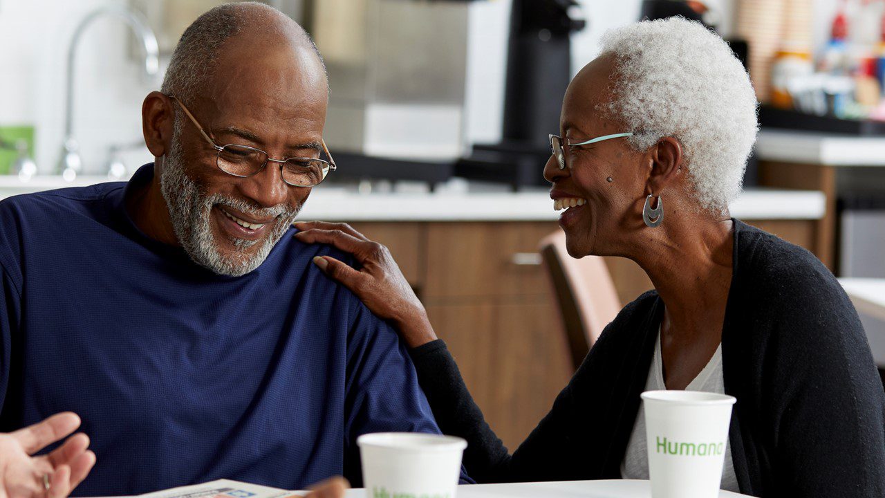 Older African American couple sitting at table smiling with visible Humana cup