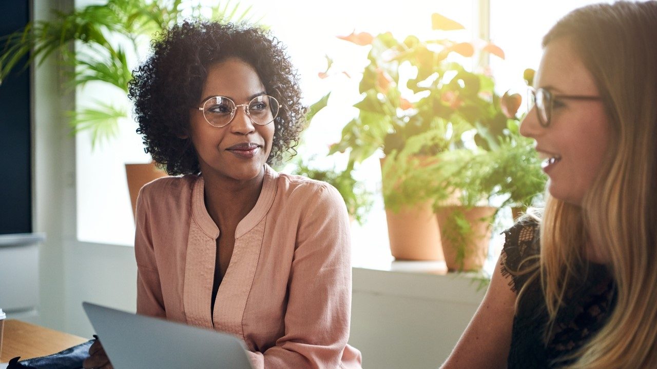 Two women working at table with computers speaking with each other