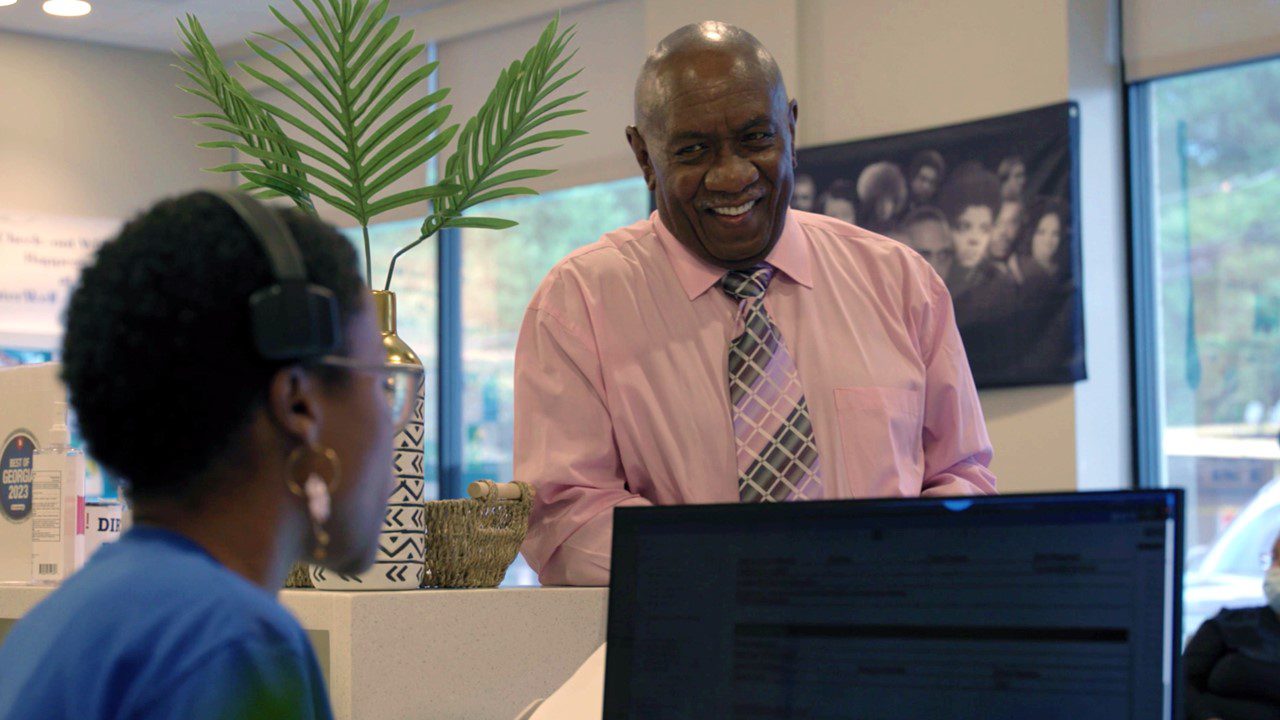 Older African American man in pink shirt talking to receptionist at health clinic