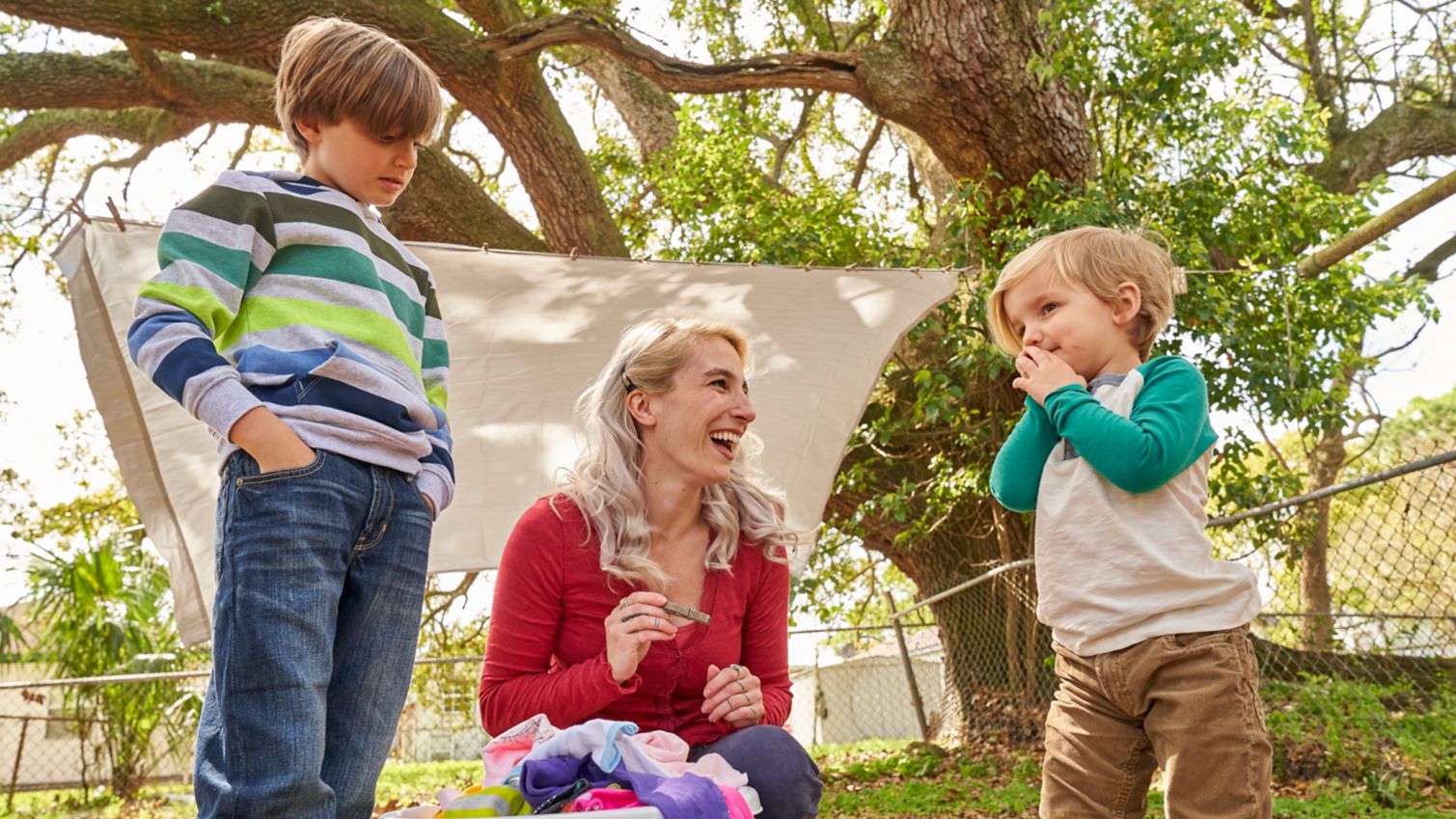 Mother and her two sons outside pulling laundry off the line and placing it in the hamper on the ground.