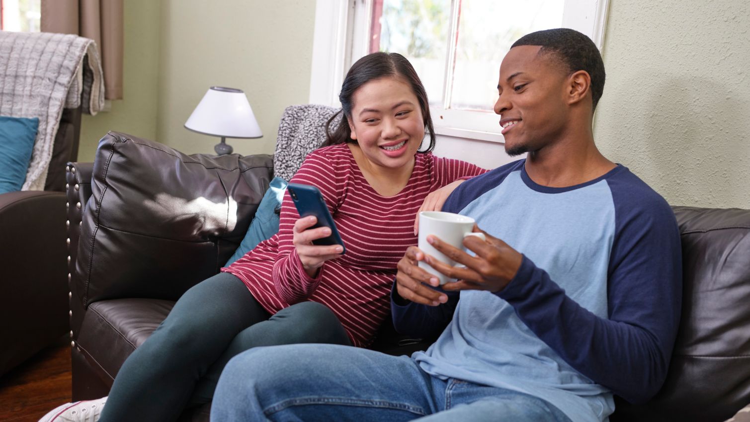 A smiling couple sits on the couch and reads an article on a cell phone together