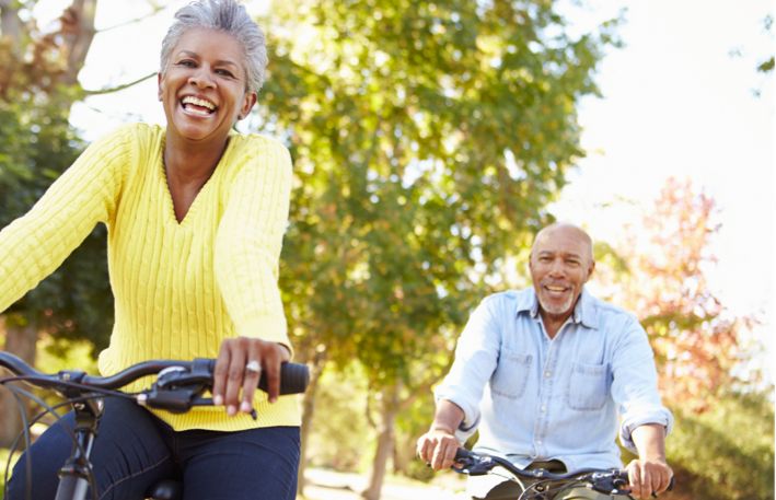 On a bright summer morning an athletic older couple riding bicycles in the open with trees behind them.