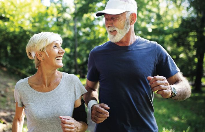 On a bright summer morning an athletic older couple smiling at each other and running out in the open with trees behind them.