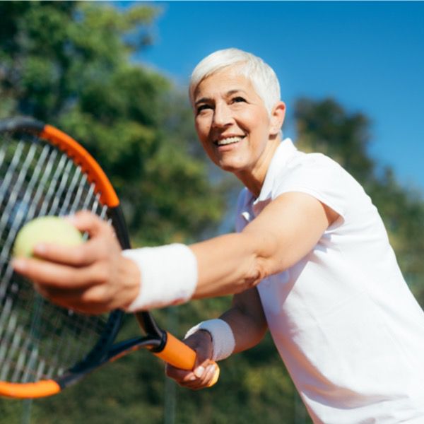 An athletic older woman about to serve a tennis ball out in the open with blue sky and green trees in the background.