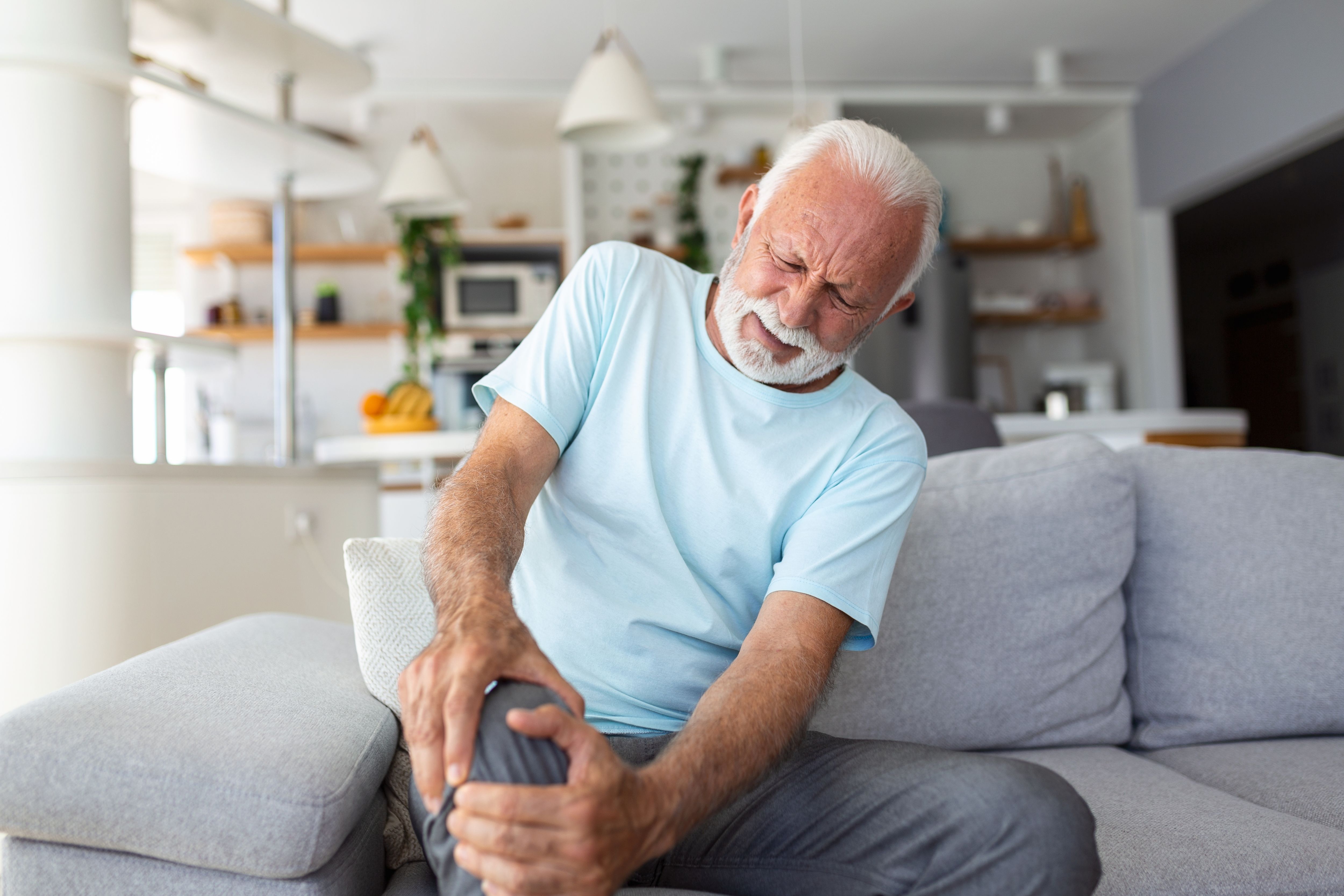 The older man is sitting on the couch at home, has pain in the knee joint, holding his leg, osteoarthritis concept.
