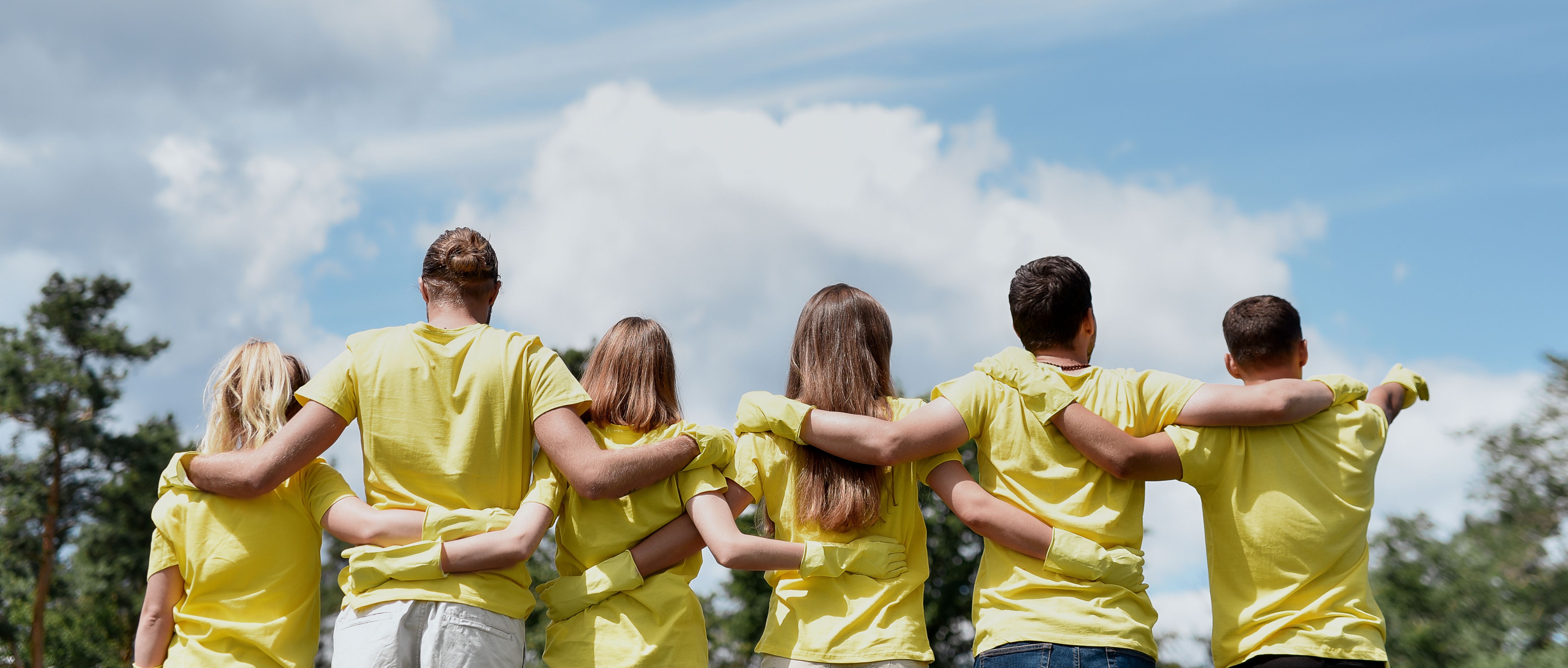 Love your nature. Group of young volunteers wearing uniform and rubber gloves hugging and looking at green forest in front of them, rear view. Environment and ecology concept, charity work